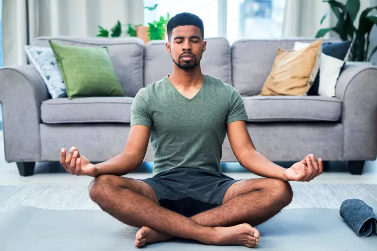 Homem negro sentado num tapete cinza na sala fazendo meditação